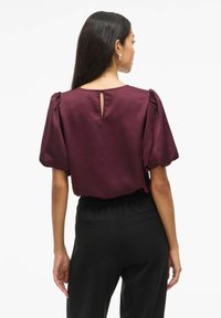 Top in satin bordeaux con maniche a sbuffo, dettaglio a chiave sul retro, abbinato a pantaloni neri a vita alta con tasche laterali.