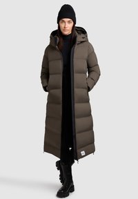 Vrouw in een lange, gewatteerde bruine jas met capuchon, zwarte beanie, zwarte coltrui, zwarte broek en chunky zwarte laarzen, staand tegen een neutrale achtergrond.
