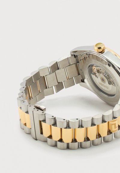 Dviejų tonų metalinė laikrodžio apyrankė su sidabrinėmis ir auksinėmis grandelėmis bei permatoma nugarėle, atskleidžiančia vidines mechanizmas ir "Versace" ženklinimą.