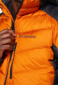 Ruka osoby upravuje zip na oranžové bundě Columbia s výplní, což ukazuje texturu a logo na přední straně.