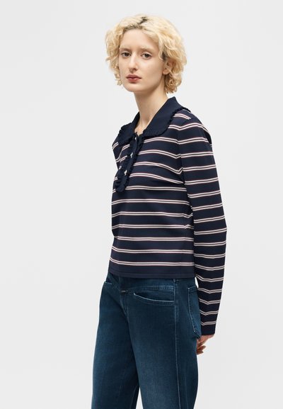 Γυναίκα με κοντά σγουρά ξανθά μαλλιά που φοράει μπλε μακρυμάνικο ριγέ πουκάμισο με γιακά με βολάν και σκούρο μπλε τζιν, στέκεται πλάγια.