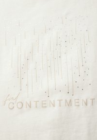 Bílá tkanina s třpytivými stříbrnými a zlatými akcenty tvořícími vzor připomínající déšť. Text zní "najdi SPOKOJENOST" v jemném písmu.