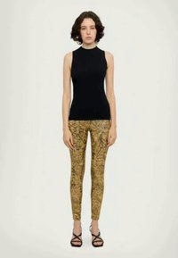 Juodas be rankovių viršus, derinamas su auksinėmis raštuotomis legginsomis; modelis vilki juodas šlepetes su dirželiais, stovintis prieš paprastą šviesią foną.