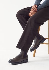 Braune Leder-Stiefeletten mit Brogue-Details, elastischen Seitenteilen und strukturierten Sohlen, kombiniert mit dunklen Hosen auf einem Holzstuhl.