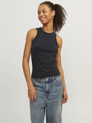 Ung kvinne med krøllete hår som har på seg svart ermeløs ribbestrikket topp og løse blå jeans, smilende og ser til siden.