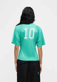 Άτομο με μακριά σκούρα μαλλιά στέκεται με την πλάτη προς τα εμπρός, φορώντας μια πετρόλ κοντομάνικη μπλούζα με τον αριθμό "10" και μαύρο παντελόνι καρότσα.