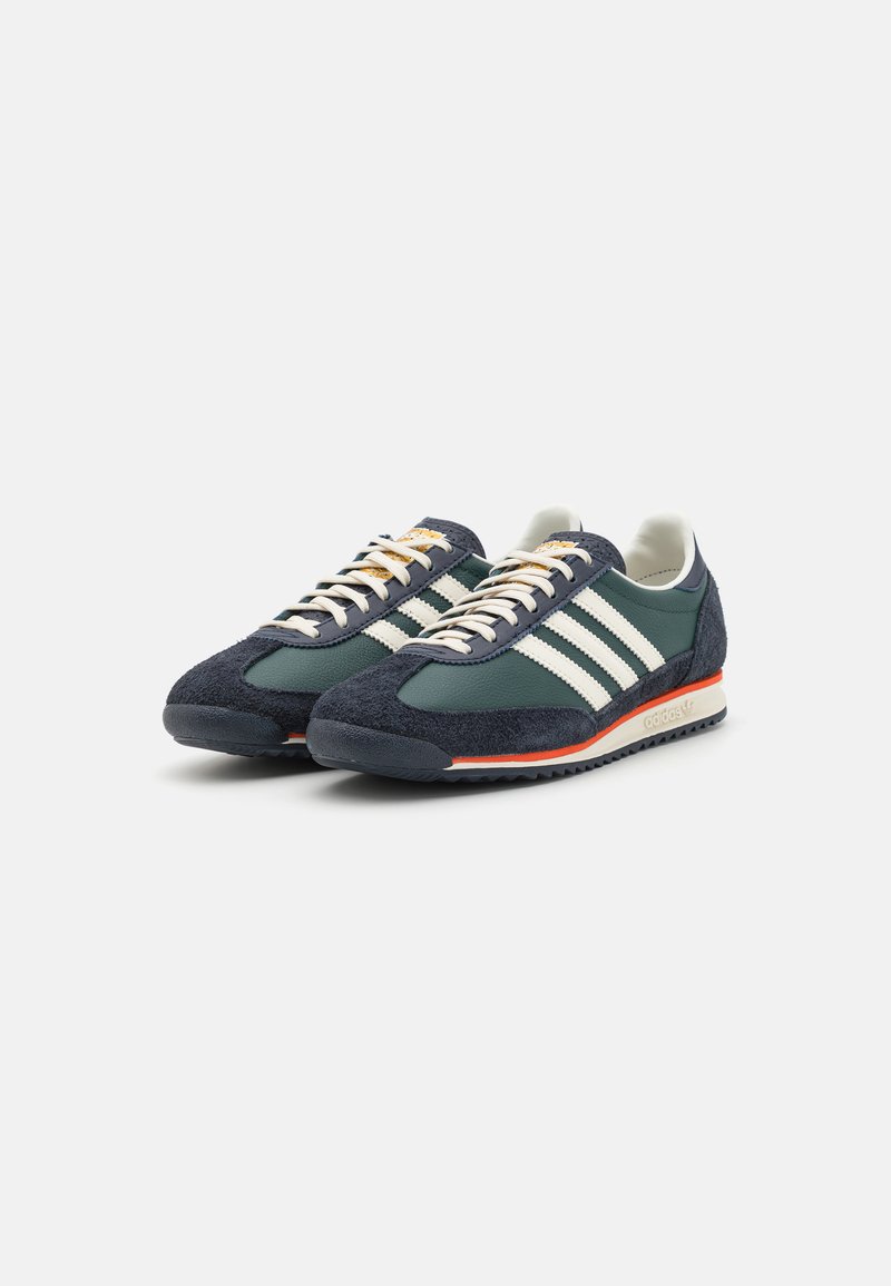 Zapatillas de ante verde y azul marino con rayas blancas, acentos en crema y detalles naranjas en la suela. Cierre con cordones y acabado texturizado.