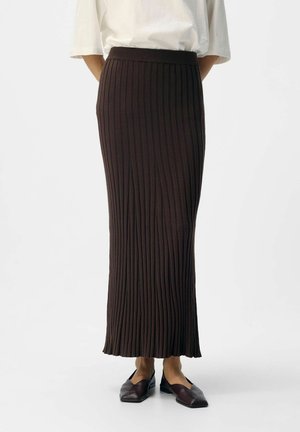 Femme portant une jupe midi en tricot côtelé marron foncé taille haute avec des ballerines noires et un haut blanc à manches courtes rentré, debout sur un sol blanc.