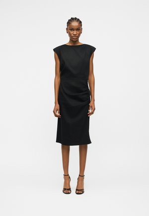 Γυναίκα που στέκεται φορώντας μαύρο μίντι φόρεμα χωρίς μανίκια με ελαφριά πτυχοποίηση στο πλάι, και μαύρα ψηλοτάκουνα σανδάλια με λουράκια, μπροστά σε λευκό φόντο.