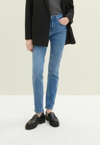 Personne portant un jean slim bleu, des mocassins noirs, un blazer noir et un haut foncé, debout avec une main dans la poche sur fond clair.