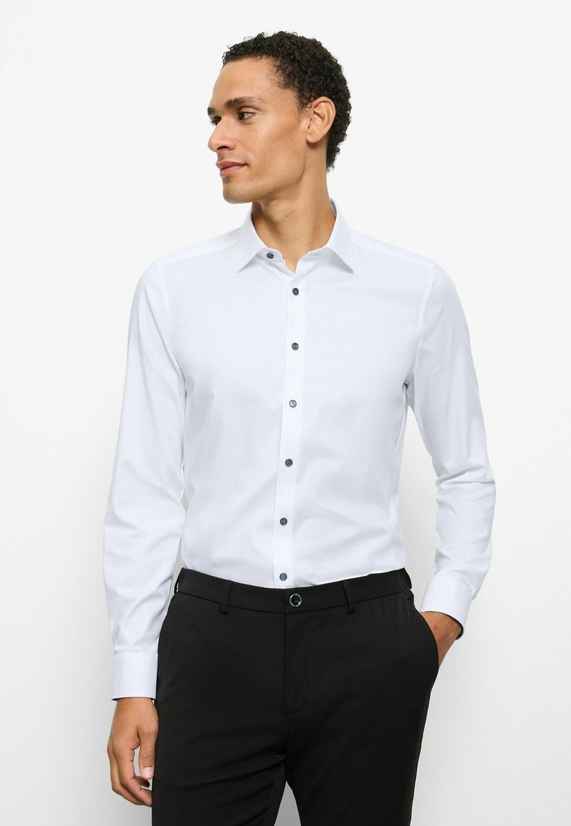 White long-sleeve shirt with a button-down collar, featuring black buttons down the front. Smooth fabric, paired with black trousers.