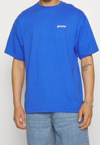 Muž oblečený v královsky modrém tričku Dickies s malým logem na hrudi, světle modrých džínách, stříbrném řetízkovém náramku a tetováním na pravém předloktí.