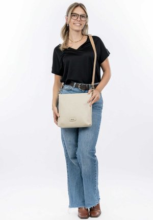 Beige leather crossbody bag with a textured finish, adjustable strap, silver hardware, and a front logo detail. Casual outfit with black top and blue jeans.