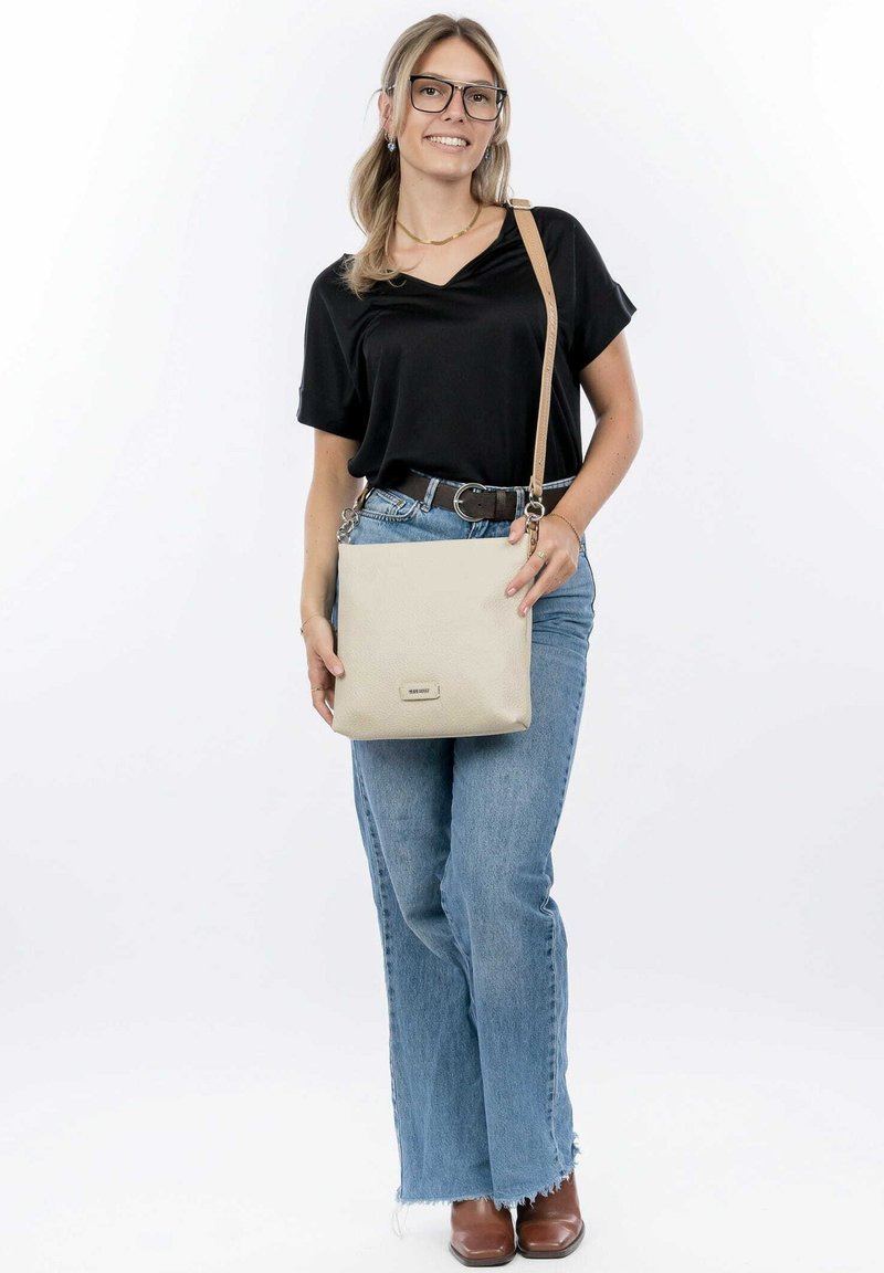 Borsa a tracolla in pelle beige con finitura testurizzata, tracolla regolabile, accessori in argento e dettaglio del logo frontale. Outfit casual con top nero e jeans blu.