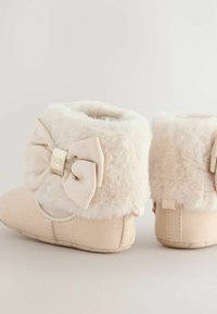 Weiche, beige Baby-Schühchen mit flauschigen Bündchen und dekorativen Stoffschleifen an den Seiten vor einem neutralen Hintergrund.