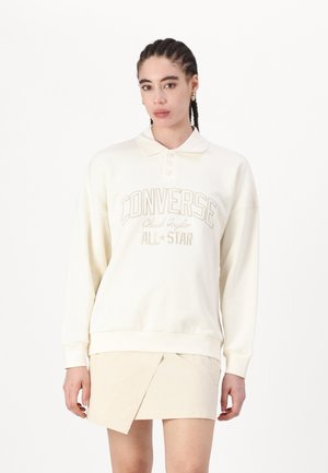 Sudadera color crema con cuello de polo, logo bordado de "Converse All-Star" y puños acanalados. Combinada con una falda envolvente beige.