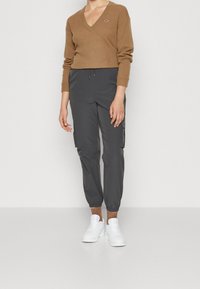 Ruskea V-päähine neulepaita, joka on valmistettu pehmeästä materiaalista, yhdistetty tummanharmaisiin kapeisiin cargo-housuihin. Valkoiset lenkkarit täydentävät asua.