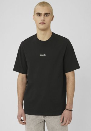 Jeune personne aux cheveux blonds courts, portant un t-shirt noir avec le texte blanc « sounds » et un jean clair, debout devant un fond uni.