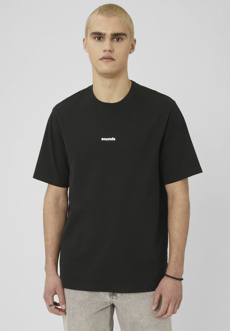 Jeune personne aux cheveux blonds courts, portant un t-shirt noir avec le texte blanc « sounds » et un jean clair, debout devant un fond uni.