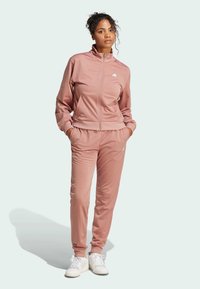 Rosa treningsoverall laget av glatt stoff; har en zip-up jakke med h�øy krage og bukser med elastisk midje. Hvite sneakers fullfører utseendet.
