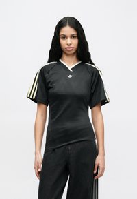 Fiatal nő hosszú fekete hajjal, fekete Adidas pólót visel, melynek ujján sárga és fehér csíkok vannak, egyszínű háttér előtt áll.