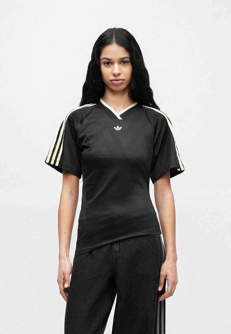 Junge Frau mit langen schwarzen Haaren, die ein schwarzes Adidas-Shirt mit gelben und weißen Streifen auf den Ärmeln trägt, vor einem einfarbigen Hintergrund stehend.