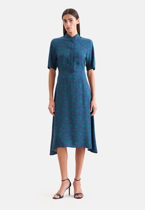 Kobieta ubrana w niebieską sukienkę midi z zielonym kwiatowym nadrukiem, krótkimi rękawami, wysokim kołnierzem i czarnymi szpilkami z paskiem wokół kostki, stojąca na białym tle.