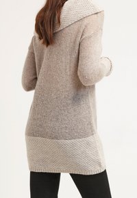 Long cardigan en tricot avec un dégradé beige clair. Présente une grande capuche texturée, des poignets côtelés et une bande solide plus claire au niveau de l'ourlet.