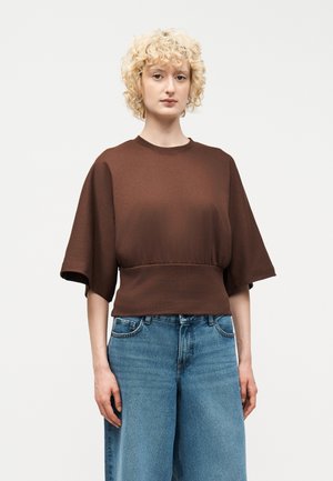 Γυναίκα με σγουρά ξανθά μαλλιά, φορώντας φαρδιά καφέ μπλούζα με φαρδιά μανίκια και ψηλόμεσο μπλε τζιν, στέκεται μπροστά σε απλό φόντο.