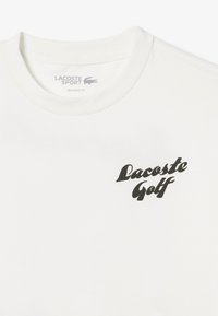 Camiseta blanca de algodón con corte relajado, que presenta un logo negro de "Lacoste Golf" en el lado izquierdo y un escote redondo.
