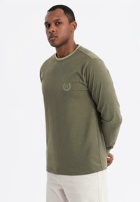 Uomo che indossa una camicia a maniche lunghe verde oliva con un piccolo logo "EX" sul petto e pantaloni chiari, in piedi con le mani dietro la schiena.