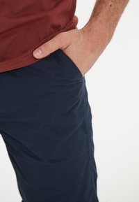 Marineblaue Hose mit leichtem Glanz, ausgestattet mit einer Seitentasche und einem entspannten Schnitt, kombiniert mit einem bordeauxfarbenen Rundhalsshirt.