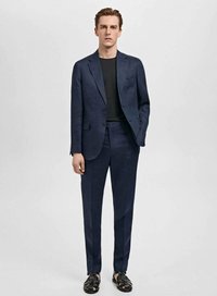 Costume bleu marine avec une veste cintrée et un pantalon slim, fabriqué en tissu texturé. Porté par-dessus une chemise noire, assorti à des sandales noires.