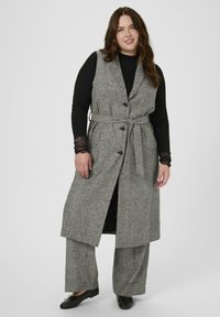 Harmaa houndstooth-mallinen pitkä liivi, jossa on etunapit ja vyö. Yhdistetty mustan pitkän hihansuun paidan ja harmaiden leveälahkeisten housujen kanssa. Sulava kangas.