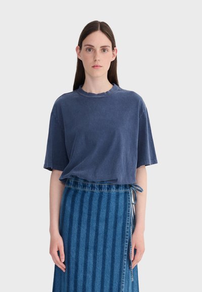 Jeune femme aux longs cheveux bruns portant un t-shirt bleu marine délavé et ample ainsi qu'une jupe en denim bleu taille haute avec un lien latéral.