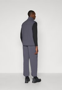 Sleeveless, dark grey vest over black long-sleeve shirt; paired with matching grey pants and black athletic shoes, viewed from behind.