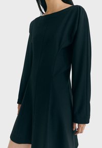 Schwarzes Langarmkleid mit Bootsausschnitt, strukturiertem Schnitt und ausgestelltem Rock, aus einem glatten Stoff mit minimalen Nähten.