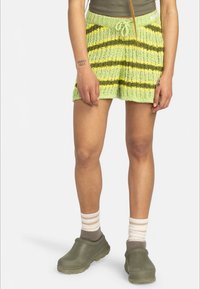 Person, der grün- und gelbgestreifte Strickshorts, olivgrüne Schlappen und weiße Socken mit beigen Streifen trägt, steht vor einem weißen Hintergrund.