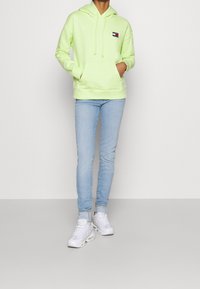 Sudadera verde neón de material suave con un bolsillo frontal, capucha con cordones y un pequeño parche de logo. Combinada con unos jeans ajustados de color azul claro y zapatillas blancas.