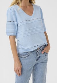 Mujer vistiendo un suéter de textura azul claro con escote en V y mangas cortas, combinado con jeans de mezclilla azul claro, con la mano en el bolsillo, frente a un fondo liso.