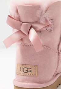 Botte en daim rose avec un intérieur doux et pelucheux. Elle présente un nœud en ruban de satin et un patch en cuir de marque sur le côté.