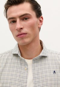 White checkered shirt with navy lines, button-down collar, and embroidered skull logo on the chest, layered over a light-colored T-shirt.