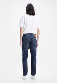 Vaqueros de denim azul con corte recto, con distintivos bolsillos traseros y una etiqueta de cuero marrón en la cintura, combinados con una camisa blanca de manga corta.