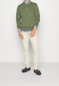 Muž ve olivově zeleném svetru přes modrou kostkovanou košili, béžových kalhotech a námořnických mokasínech s hnědými tkaničkami stojí na bílém pozadí.