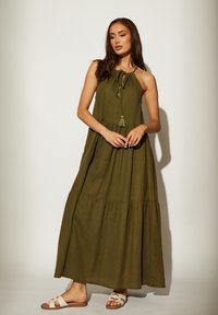 Olijfgroen maxi-jurk met een gelaagd ontwerp, halternek en franjeaccenten. Model draagt beige sandalen met banden.