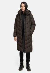 Cappotto lungo marrone imbottito con cappuccio, dotato di zip davanti, tasche laterali e motivo a rombi. Indossato sopra una maglia nera e pantaloni.