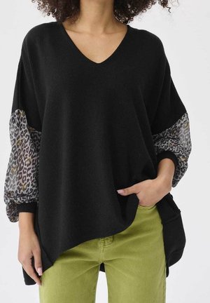 Femme portant un pull noir à col en V avec des manches ballon transparentes imprimées léopard et un pantalon vert olive, main dans la poche.