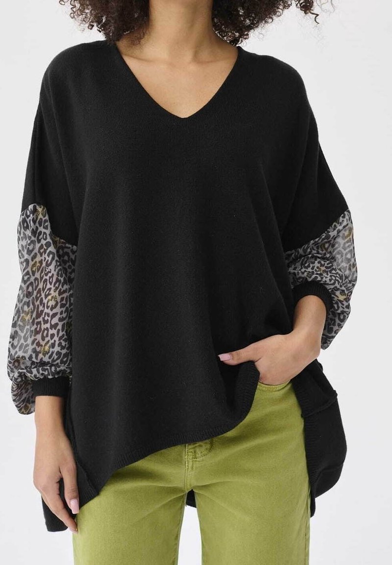 Femme portant un pull noir à col en V avec des manches ballon transparentes imprimées léopard et un pantalon vert olive, main dans la poche.
