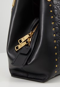 Gros plan d'un coin de sac en cuir noir avec tirettes de fermeture éclair dorées, motif embossé sur le côté et une rangée de clous dorés le long du bord.