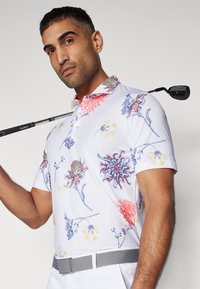 Golf polotrøye i hvit med flerfargede blomster mønstre. Har en solid krage i rød og grå belte. Teksturen ser glatt ut.